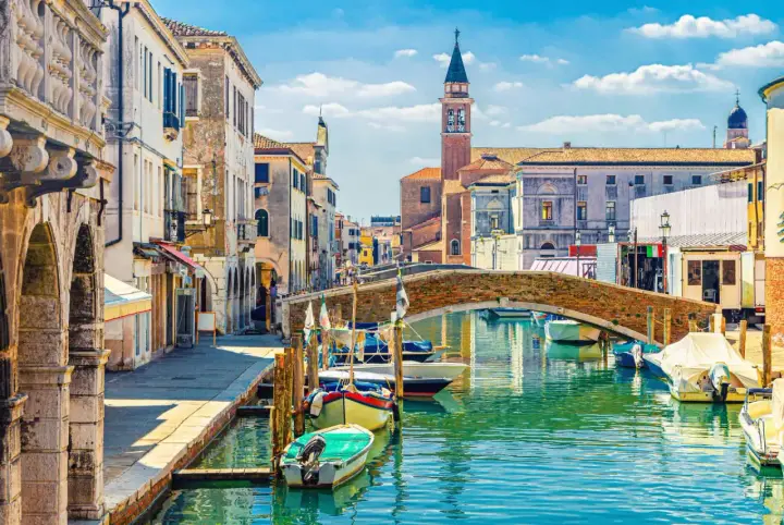 Un canal vénitien bordé de maisons colorées, avec des bateaux amarrés et un pont de briques. L'envie d'aller à Venise est forte.